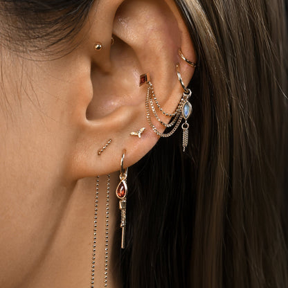 Close up ear styling of 14K gold charm of a a marquise-cut natural gemstones paired with three delicate gold curb chain tassels along with chain, hoop, threader and stud items