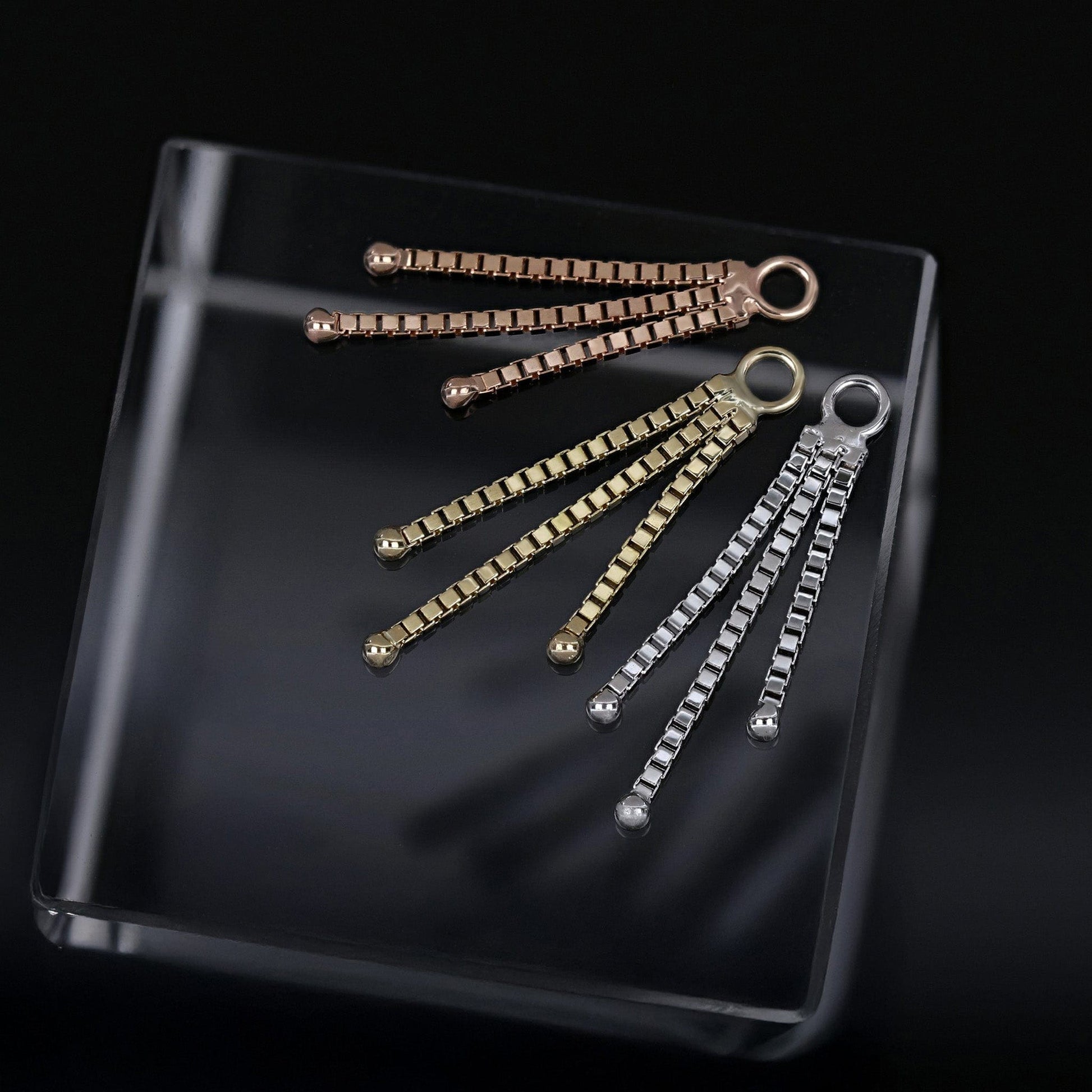 Rose, yellow, and white gold chain charms on a clear cube, set against a dark background